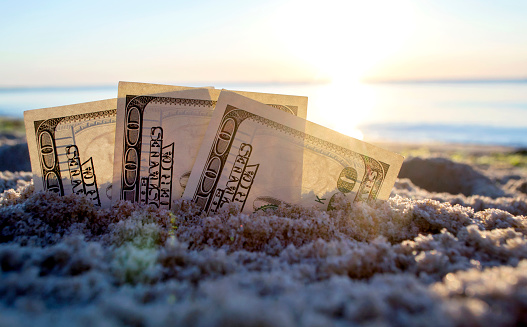 Three dollar bills are buried in sand on sandy beach near sea at sunset dawn in summer close-up. Dollar bills partially buried in sand