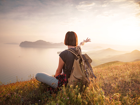 A young Asian woman with a backpack Sitting on a hill slope and enjoying the view of the sunset sea and mountains . Mountain and coastal travel, freedom and an active lifestyle