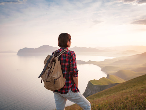 A young Asian woman with a backpack hiking in the summer and enjoying the view of the sunset sea and mountains. Mountain and coastal travel, freedom and an active lifestyle