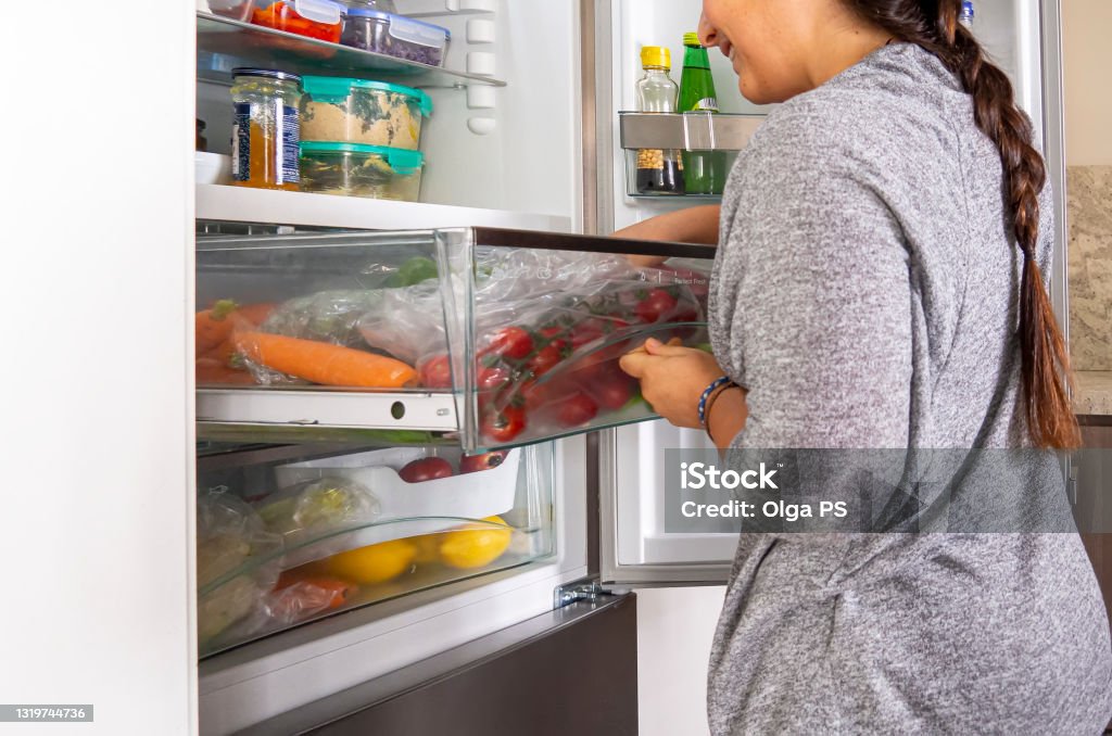 Young woman searching in a fridge draw for vegetables Young woman searching in a fridge draw for vegetables for cooking a salad. Open refrigerator drawer with tomatoes and carrots, food leftover containers on the shelves. Real vegetarian household fridge Refrigerator Stock Photo