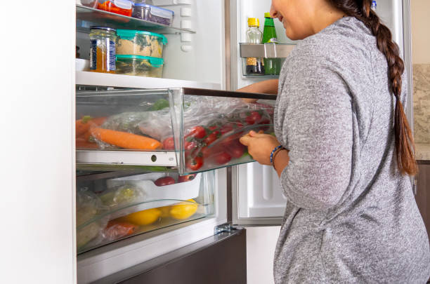 Young woman searching in a fridge draw for vegetables Young woman searching in a fridge draw for vegetables for cooking a salad. Open refrigerator drawer with tomatoes and carrots, food leftover containers on the shelves. Real vegetarian household fridge drawer fridge stock pictures, royalty-free photos & images