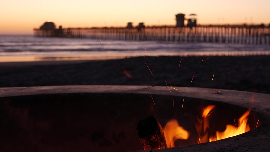 Campfire pit by Oceanside pier, California USA. Camp fire burning on ocean beach, bonfire flame in cement ring place for bbq, sea water waves. Romantic evening twilight sky, dusk after summer sunset.