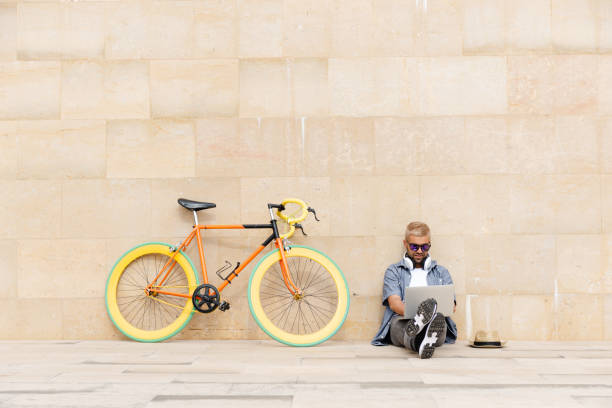 Hispanic man with a colorful urban bike. Hispanic hipster freelancer sitting on the floor against the wall and working on his laptop beside an urban colorful bicycle. digital nomad stock pictures, royalty-free photos & images
