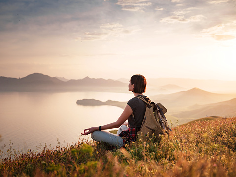 A young Asian woman with a backpack Sitting on a hill slope and enjoying the view of the sunset sea and mountains and meditates with a beautiful view .