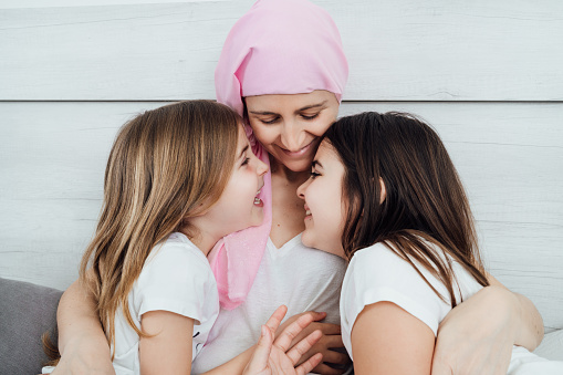 A mother with cancer wears a pink headscarf, tenderly and happily hugging her two blond and brown-haired daughters. They are sitting on the bed with a white background