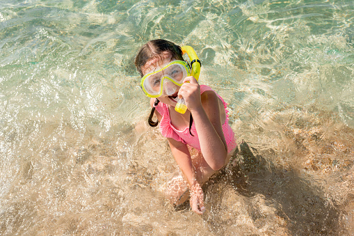Top view of girl in pink swimsuit with snorkeling mask sitting on sandy beach. Child is ready for diving. Happy kid with goggles and snorkel on summer vacation.