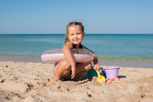 Portrait of cheerful child girl playing with sand at the beach in summer day. Kids building sand sculptures with shovels and buckets on coast sea. Fun family vacation concept