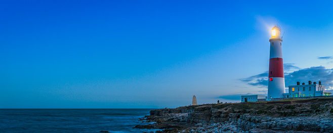 Lighthouse shining its beams across the cliffs of a rocky peninsula out to the dark ocean beyond as the warm light of sunset fades in the panoramic skies above, Portland Bill, Dorset. UK.
