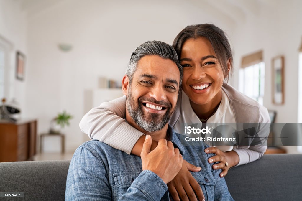 Mature indian couple hugging and looking at camera Portrait of multiethnic couple embracing and looking at camera sitting on sofa. Smiling african american woman hugging mid adult man sitting on couch from behind at home. Happy mature mixed race couple laughing at home. Couple - Relationship Stock Photo
