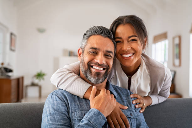 Mature indian couple hugging and looking at camera Portrait of multiethnic couple embracing and looking at camera sitting on sofa. Smiling african american woman hugging mid adult man sitting on couch from behind at home. Happy mature mixed race couple laughing at home. couple relationship stock pictures, royalty-free photos & images