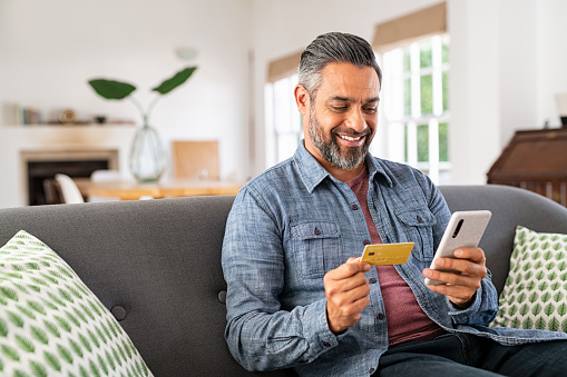 Middle eastern mature man with beard using credit card to make online payment on smartphone. Mixed race man holding debit card and using cellphone for shopping online from home. Smiling indian guy using smart phone to check credit card transactions from app.