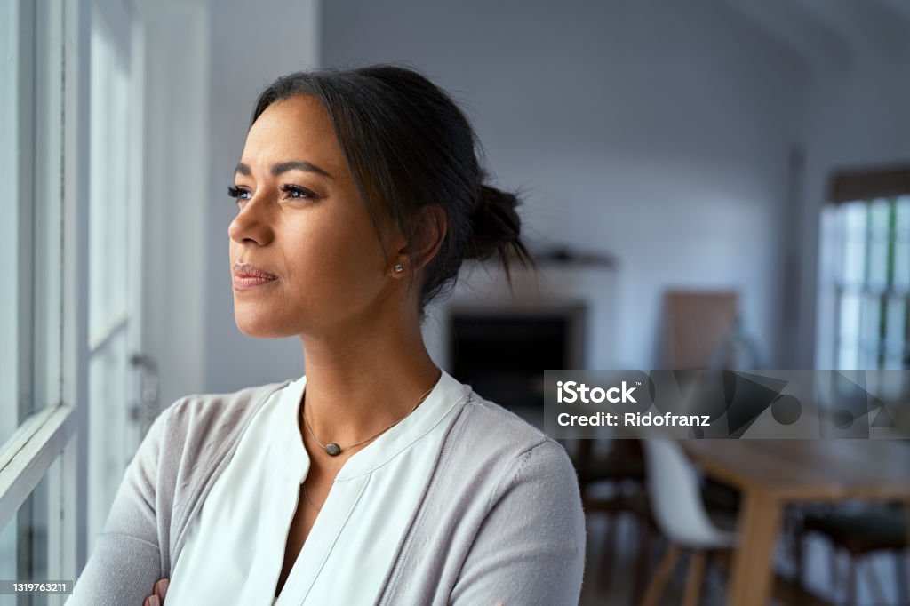 Thoughtful black woman looking outside window Mature african woman looking outside window with uncertainty. Thoughtful mid adult woman looking away through the window while thinking about her future business after pandemic. Close up face of doubtful lady at home with pensive expression and copy space. Contemplation Stock Photo