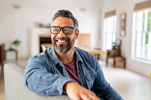 Happy mature middle eastern man wearing eyeglasses with beard sitting on couch at home. Portrait of indian man relaxing at home and looking away with big smile. Handsome mid adult guy with specs thinking about his future.