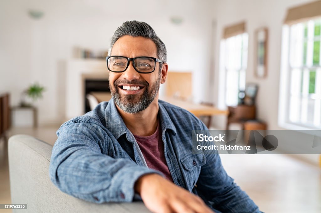 Mature ethnic man wearing eyeglasses at home Happy mature middle eastern man wearing eyeglasses with beard sitting on couch at home. Portrait of indian man relaxing at home and looking away with big smile. Handsome mid adult guy with specs thinking about his future. Men Stock Photo