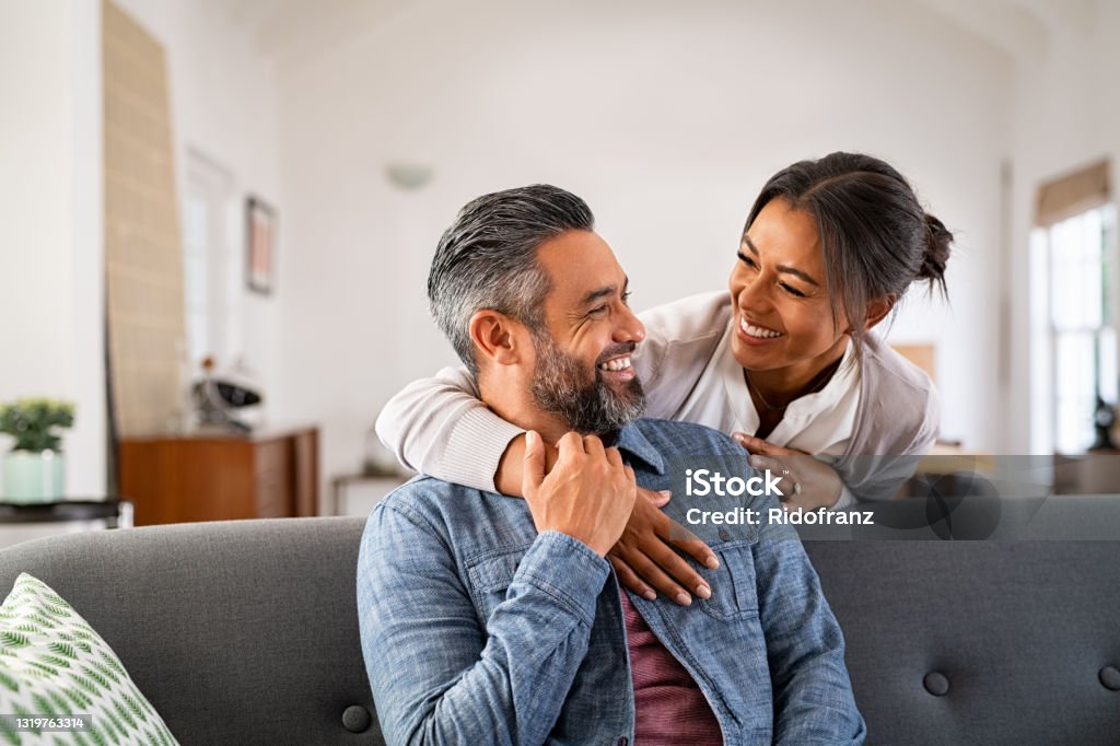 Mature multiethnic couple laughing and embracing at home Smiling ethnic woman hugging her husband on the couch from behind in the living room. Middle eastern man having fun with his beautiful young wife on the couch. MId adult indian man with latin woman laughing and looking at each other at home: complicity and love concept. Couple - Relationship Stock Photo