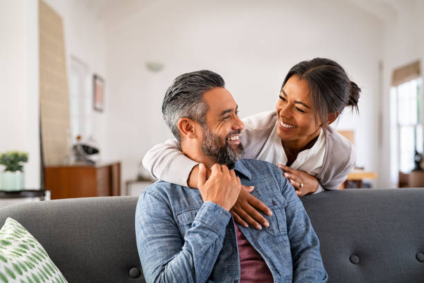 Mature multiethnic couple laughing and embracing at home Smiling ethnic woman hugging her husband on the couch from behind in the living room. Middle eastern man having fun with his beautiful young wife on the couch. MId adult indian man with latin woman laughing and looking at each other at home: complicity and love concept. health lifestyle stock pictures, royalty-free photos & images