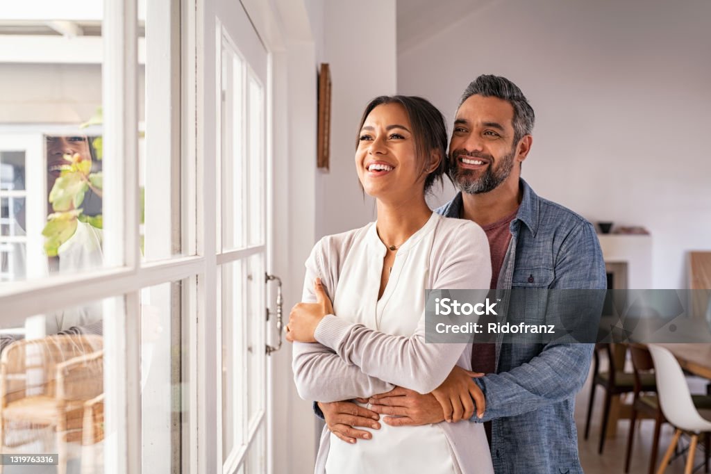 Mature multiethnic couple thinking about their future family Smiling mid adult couple hugging each other and standing near window while looking outside. Happy and romantic mature man embracing hispanic wife from behind while standing at home with copy space. Future, vision and daydream concept. Couple - Relationship Stock Photo