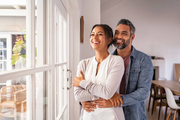 Mature multiethnic couple thinking about their future family Smiling mid adult couple hugging each other and standing near window while looking outside. Happy and romantic mature man embracing hispanic wife from behind while standing at home with copy space. Future, vision and daydream concept. indian ethnicity stock pictures, royalty-free photos & images