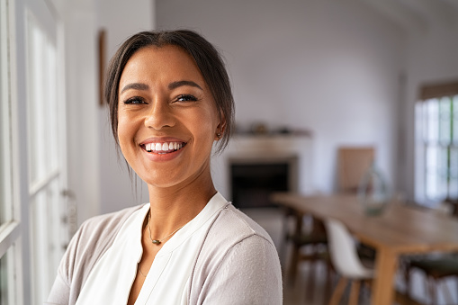 Portrait of a beautiful african woman smiling while looking at camera. Mature black woman in casual standing near window at home. Close up face of laughing cheerful lady near window with copy space.