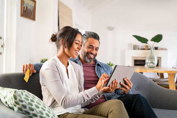 Multiethnic mid adult couple using digital tablet at home Happy multiethnic mature couple using digital tablet for online payment with credit card. Cheerful latin wife showing something to buy on digital tablet to her indian husband while holding bank card. Mid adult hispanic woman and middle eastern man sitting on couch in living room while doing shopping online together. indian woman smiling stock pictures, royalty-free photos & images
