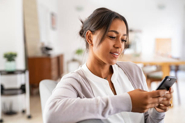 African mid woman using smartphone at home Mature african woman sitting on couch at home using smartphone. Black woman relaxing on the sofa while messaging with a cellphone at home. Happy mature indian woman typing on mobile phone in living room. house phone stock pictures, royalty-free photos & images