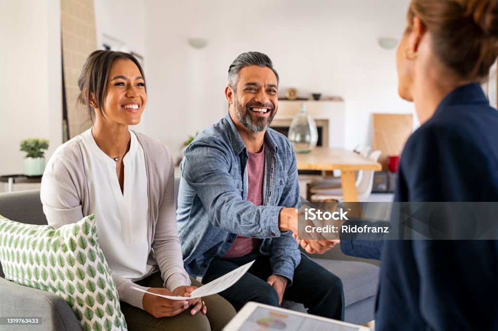 Multiethnic couple handshake with consultant at home Mature indian man shaking hands with financial advisor at home. Happy smiling couple greeting broker with handshake at home. Multiethnic mid adult man and hispanic woman sealing a contract. Customer Stock Photo
