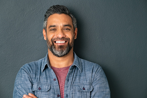 Close up face of handsome mature man with beard looking at camera. Happy mid adult indian man isolated against grey wall. Portrait of smiling mixed race guy on gray background with copy space.
