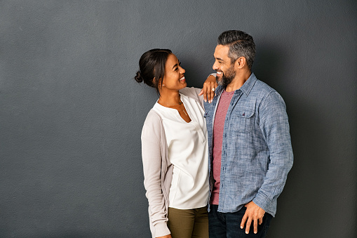 Happy mature indian couple embracing and looking at each other against gray background. Mid adult multiethnic couple in love standing against grey wall. Middle eastern man fall in love of his beautiful hispanic girlfriend.