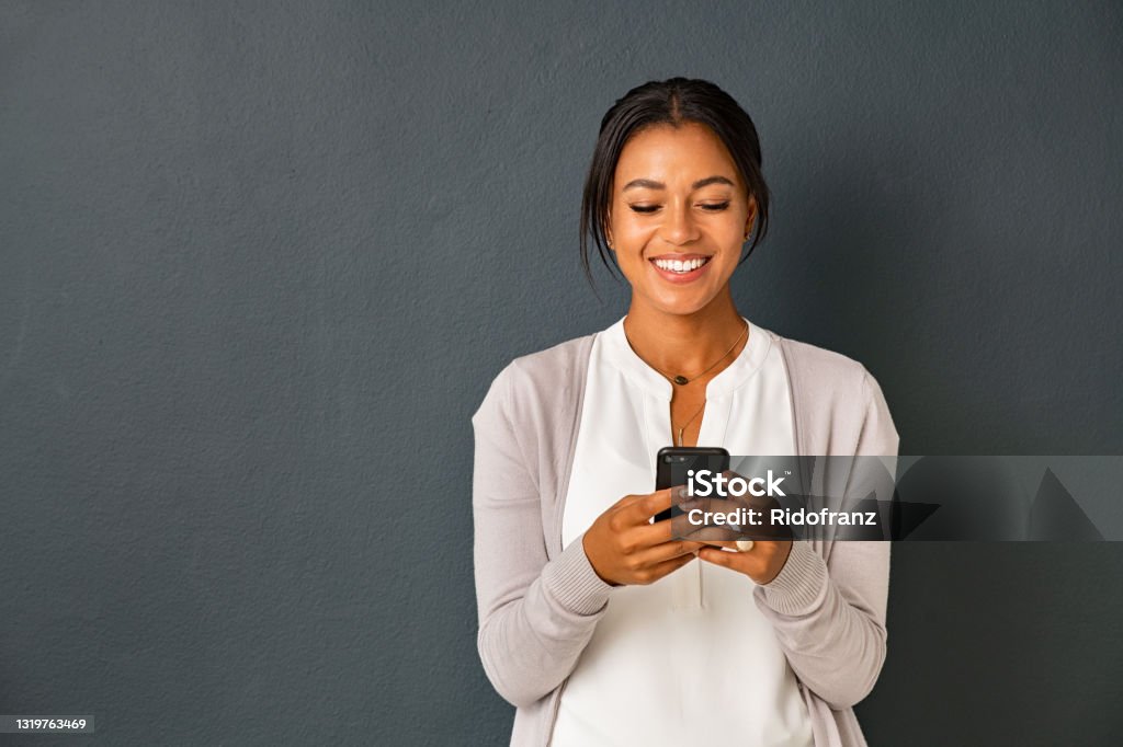 Happy african american woman using smartphone Mid adult african american woman texting message on smart phone isolated on grey background. Smiling indian woman using mobile phone and looking at it. Happy casual multiethnic lady messaging on the smartphone with new app. Women Stock Photo