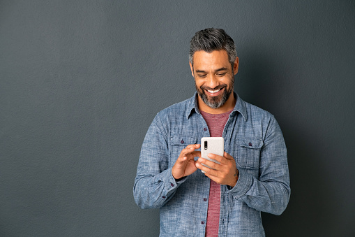 Mid adult multiethnic man texting phone message on smart phone isolated on grey background. Smiling middle eastern man using smartphone leaning on gray wall. Happy mixed race guy using new app on mobile phone with copy space.