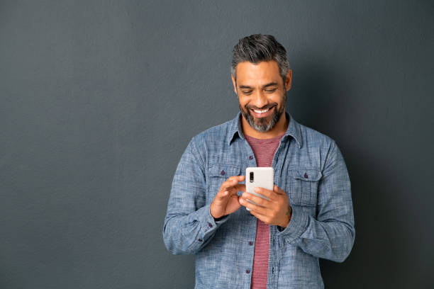 Mature indian man messaging on smartphone Mid adult multiethnic man texting phone message on smart phone isolated on grey background. Smiling middle eastern man using smartphone leaning on gray wall. Happy mixed race guy using new app on mobile phone with copy space. person on phone stock pictures, royalty-free photos & images