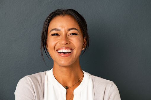 Close up face of beautiful mature woman looking at camera with big laugh. Mid adult hispanic woman standing isolated against grey wall while looking at camera. Portrait of smiling indian lady laughing on gray background.