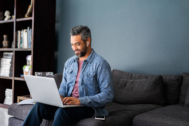 Indian man typing on laptop while working from home Successful mature indian businessman sitting on couch typing on laptop with wireless earphones. Mixed race businessman sitting on couch while working from home during video call. Happy multiethnic business man in casual clothing working on computer. working from home stock pictures, royalty-free photos & images