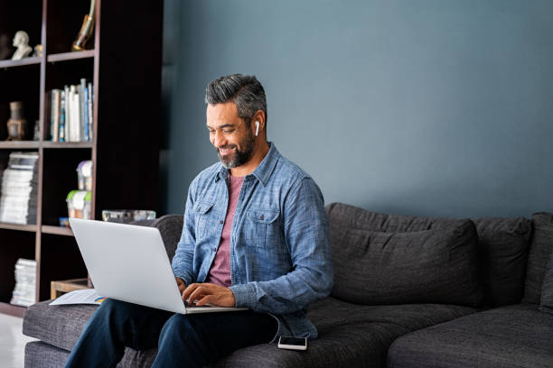 Indian man typing on laptop while working from home Successful mature indian businessman sitting on couch typing on laptop with wireless earphones. Mixed race businessman sitting on couch while working from home during video call. Happy multiethnic business man in casual clothing working on computer. asian man headphones stock pictures, royalty-free photos & images
