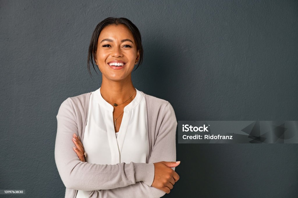 Portrait of smiling mixed race woman looking at camera Beautiful mid adult african american woman standing on grey wall with crossed arms. Mature indian woman isolated on gray background. Portrait of smiling hispanic lady looking at camera with satisfaction. Women Stock Photo