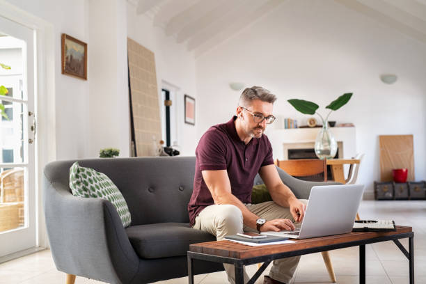 Mid adult business man working from home Mature businessman working from home on laptop with copy space. Mid adult entrepreneur focusing on work while working on laptop in living room. Manager wearing eyeglasses while working on laptop for project. mature businessman stock pictures, royalty-free photos & images