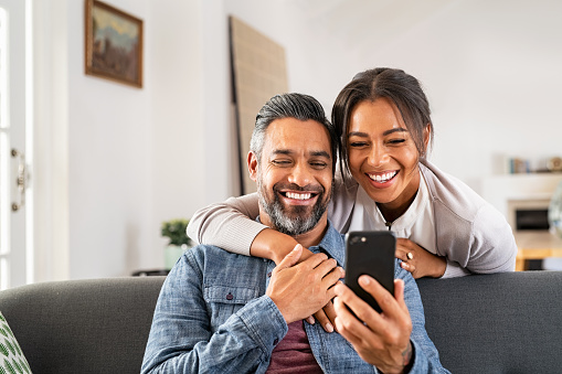 Mature multiethnic couple at home using smart phone together and smiling. Mid adult man relaxing on sofa and showing new app to african american wife on cellphone. Middle eastern man and woman sitting on couch at home and using mobile phone to do a video call with family or friends.