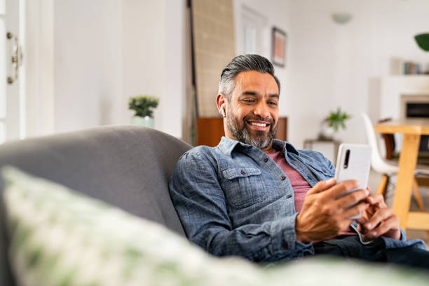 Happy mature man using smartphone while listening to music Happy mid adult man using smartphone device while sitting on sofa at home. Smiling indian man lying on couch reading messages on mobile phone while listening to music with wireless earbuds. Mature guy relaxing at home. mature men stock pictures, royalty-free photos & images