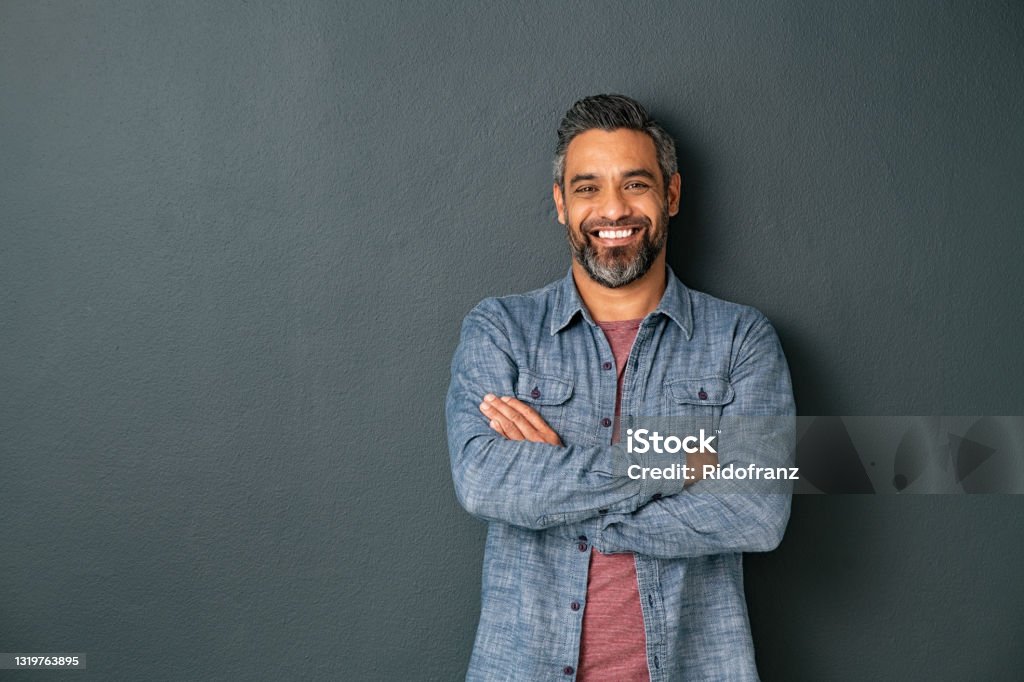 Smiling mixed race mature man on grey background Handsome mid adult man with beard standing with crossed hands and looking at camera. Mature middle eastern man isolated against grey wall and smiling. Satisfied indian guy looking at camera with a big laugh. Men Stock Photo