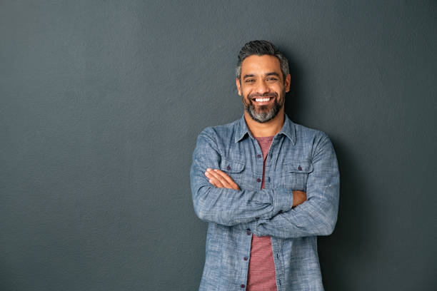 Smiling mixed race mature man on grey background Handsome mid adult man with beard standing with crossed hands and looking at camera. Mature middle eastern man isolated against grey wall and smiling. Satisfied indian guy looking at camera with a big laugh. arms crossed photos stock pictures, royalty-free photos & images