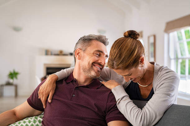 Laughing mature woman embracing man and having fun together Happy mature couple embracing on sofa while laughing and relaxing at home. Middle aged woman hugging husband from behind while smiling and joke at home. Beautiful wife and mid adult man having fun and enjoying time together. 40 49 years stock pictures, royalty-free photos & images