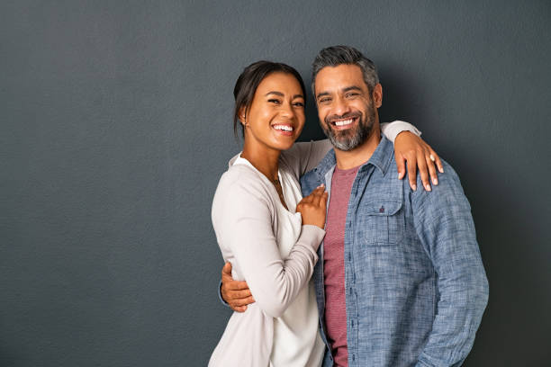 Mature multiethnic couple embracing and smiling together Portrait of happy mid adult couple embracing and looking at camera standing against gray background. Mature indian man in love standing on grey wall while hugging his beautiful hispanic woman. Portarit of carefree mixed race couple with copy space wife stock pictures, royalty-free photos & images