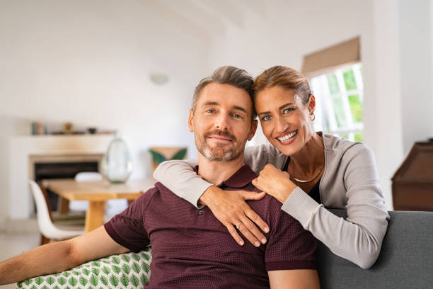 Mature woman hugging man from behind at home Middle aged couple embracing on sofa while looking at camera. Mature happy woman embracing her husband from behind while relaxing at home. Portrait of smiling wife and man loving in perfect harmony. 40 49 years stock pictures, royalty-free photos & images