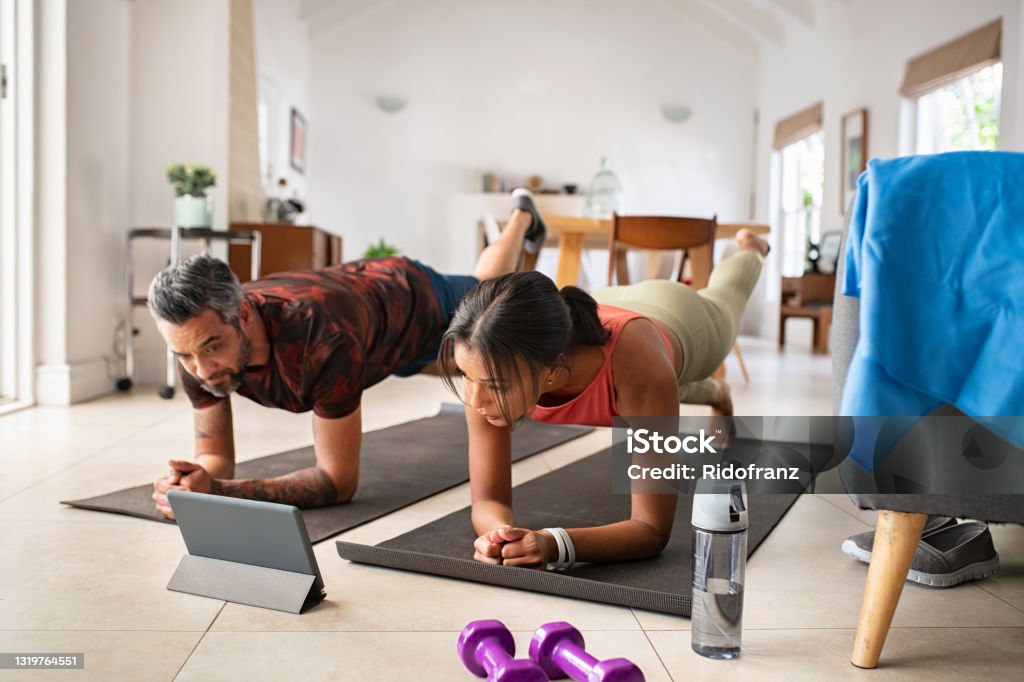 Mixed race couple practicing stretching exercise at home Multiethnic mature couple exercising at home and watching training videos on digital tablet. Middle aged woman and indian man doing planks with a leg outstretched while watching fitness lessons online on digital tablet. Fit mid adult couple doing strenght plank following online tutorials. Exercising Stock Photo