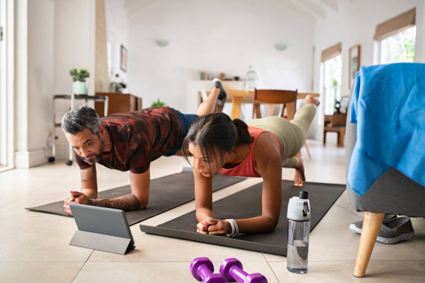 Mixed race couple practicing stretching exercise at home Multiethnic mature couple exercising at home and watching training videos on digital tablet. Middle aged woman and indian man doing planks with a leg outstretched while watching fitness lessons online on digital tablet. Fit mid adult couple doing strenght plank following online tutorials. pilates photos stock pictures, royalty-free photos & images