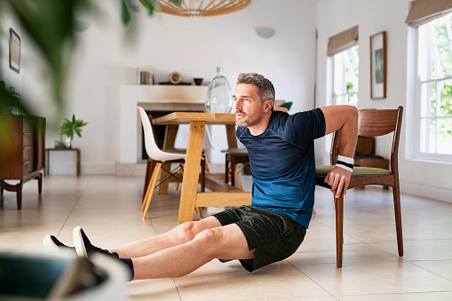 Mature man doing fitness exercise while doing pushups using chair in living room. Middle aged man training at home wearing sportswear during fitness exercises for triceps. Mid adult fit guy exercising for wellness in his living room.