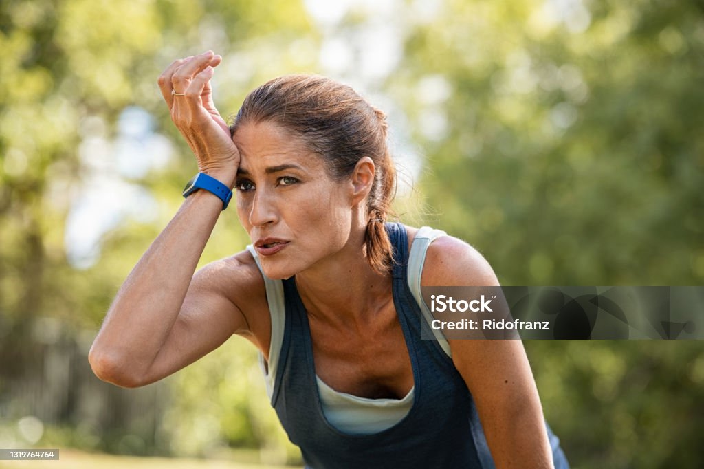 Tired mature runner wiping sweat after workout at park Exhausted mature woman after jogging catching her breath. Sweaty middle aged athlete taking a break from running at park with copy space. Tired mid adult woman wipes wet forehead after intense fitness training. Exercising Stock Photo