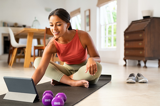 Mature healthy woman using app on digital tablet at home to train in hata yoga. Mature african american fit woman using digital tablet while sitting on yoga mat after fitness exercises. Happy smiling indian lady watching exercise tutorials on digital tablet while working out with dumbbells at home.