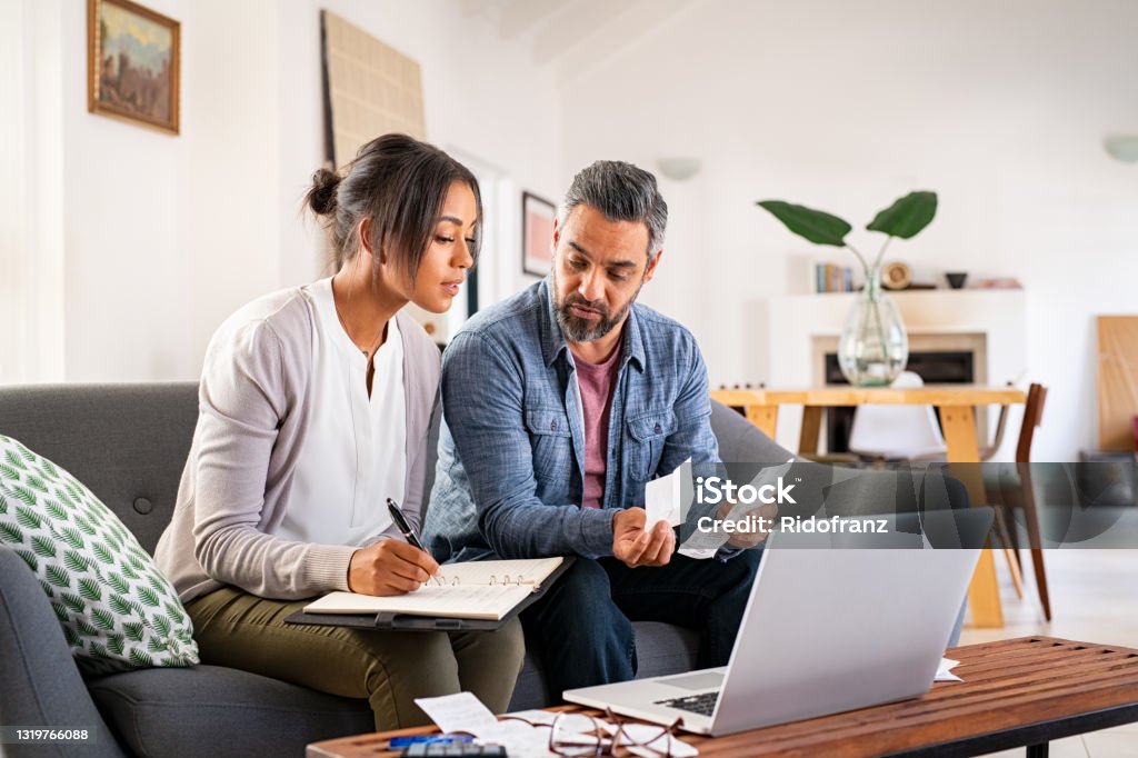 Mid adult couple working on home finance Mature couple calculating bills at home using laptop and calculator. Multiethnic couple working on computer while calculating finances sitting on couch. Mature indian man with african american woman at home analyzing their finance with documents. Finance Stock Photo