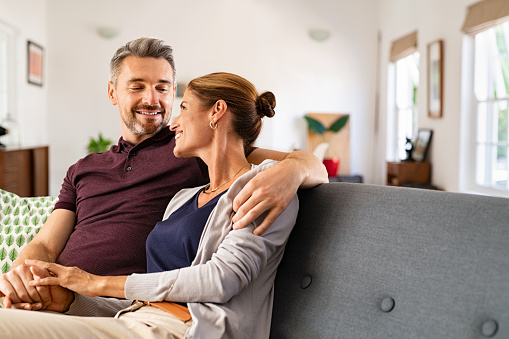 Mid adult couple embracing and relaxing on couch at home while holding hands. Romantic husband and beautiful wife hugging and looking at each other on sofa. Middle aged man relaxing at home with smiling woman.